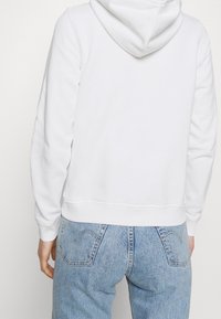 White cotton hoodie with a hood, ribbed cuffs, and hem. Worn with light blue denim jeans featuring back pockets and a faded texture.