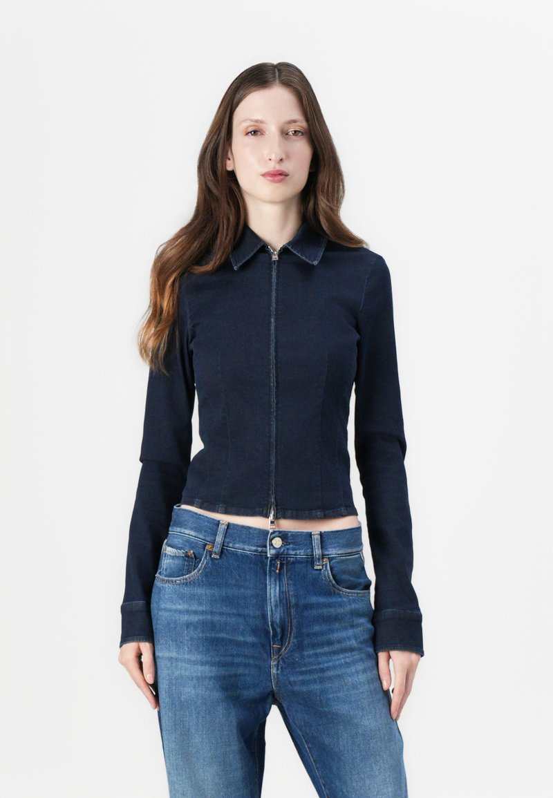 Marineblå denimjakke med krage og frontglidelås, med lange ermer og en tettsittende design, kombinert med lyseblå jeans.