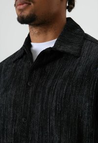 Zwart corduroy overhemd met verticale geribbelde textuur, knoopsluiting en een puntige kraag. Draagt over een wit T-shirt.