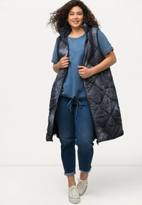 Langer, gesteppter Weste in Dunkelblau und Schwarz mit Kapuze, ausgestattet mit einem Reißverschluss, Seitentaschen und einer weich strukturierten Oberfläche. Kombiniert mit einem blauen T-Shirt und Jeans.