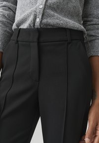 Pantaloni neri su misura con davanti piatto, texture liscia, tasche laterali e pieghe sottili, abbinati a un cardigan grigio in maglia testurizzata.