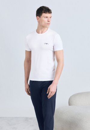 Álló férfi, szűk fehér pólót visel, amelyen apró logó látható a mellkasán, illetve sötét nadrágot, és balra néz, egyszínű háttér előtt.