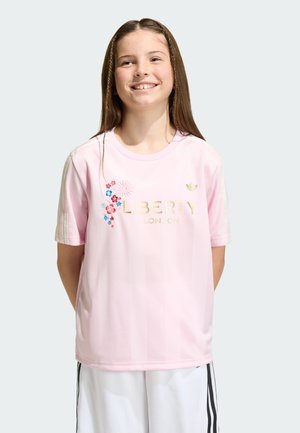 Smiling girl with long brown hair wearing a pink Liberty London t-shirt with floral embroidery and white pants with black stripes.