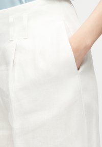 Χέρι τοποθετημένο στην μπροστινή τσέπη λευκών λινών παντελονιών με ψηλή μέση, θηλιές για ζώνη και πιέτες.