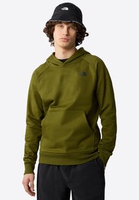 Felpa con cappuccio verde oliva con tasca a marsupio, caratterizzata da un logo nero sul petto. Abbinata a un cappello a secchiello nero.