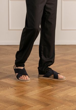 Persoon draagt zwarte broek en zwarte suède sandalen met gekruiste bandjes, staand op een houten parketvloer binnen.