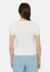 Camiseta de algodón blanca con mangas cortas, diseño ajustado y escote redondo. Llevada con pantalones vaqueros de denim azul claro que tienen bolsillos traseros.
