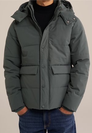 Eine Person trägt eine dunkelgrüne Daunenjacke mit Kapuze und einen schwarzen Pullover darunter, die Hände in den Taschen, vor einem hellen Hintergrund.