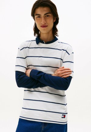 Weißes Poloshirt mit blauen Streifen, marineblauem Kragen und Ärmeln. Verfügt über ein Markenlogo-Tag am Saum. Der Stoff scheint weich und strukturiert zu sein.