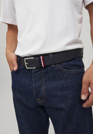 Cinturón negro con hebilla de metal plateado, con un acento a rayas rojas y blancas. Lleva puesto sobre unos jeans de mezclilla azul y una camiseta blanca.