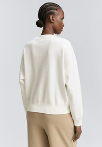 Ženska z pletenico v bunki, oblečena v ohlapen bel dolg rokav in bež hlače, obrnjena bočno proti enobarvni ozadju.