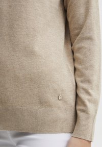 Beige kötött pulóver bordás mandzsettákkal és aljjal. A legalsó részen egy kis fémes logó részlet található, ami puha textúrát és diszkrét mintázatot展示.