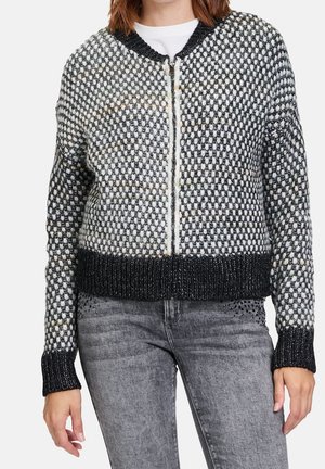 Kobieta ubrana w czarno-biały, wzorzysty, dziergany kardigan na zamek z prążkowanymi mankietami i dołem, w połączeniu z szarymi, wyblakłymi dżinsami.