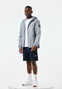 Light gray zip-up jacket with a hood, elastic cuffs, and patch on the sleeve, paired with navy cargo shorts and white sneakers.