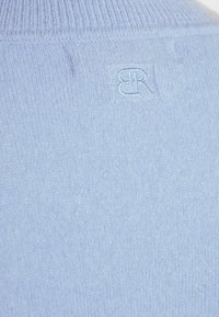 Ljusblå stickad tröja med ribbad krage. Har en distinkt broderad logotyp i nacken. Mjuk textur, slät yta.