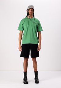 Grøn bomulds t-shirt med korte ærmer og sorte denim shorts. Båret med sorte ankelstøvler og en grå cap med dekorative detaljer.