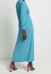 Dlouhé tyrkysové šaty s úzkým pasem, přiléhavými rukávy a hladkou texturou. V kombinaci s růžovými lodičkami s výrazným designem jehlových podpatků.