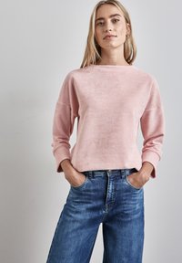 Helles rosa geripptes Sweatshirt mit weitem Bootsausschnitt und langen Ärmeln, kombiniert mit blauen hochgeschnittenen Jeans, Hände in den Taschen.