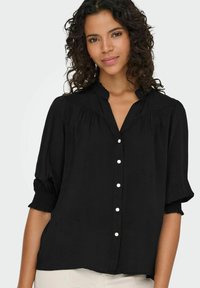 Sort bluse lavet af letvægtsstof, med en mandarin-krave, knaplukning foran og korte puffede ærmer med elastiske manchetter.