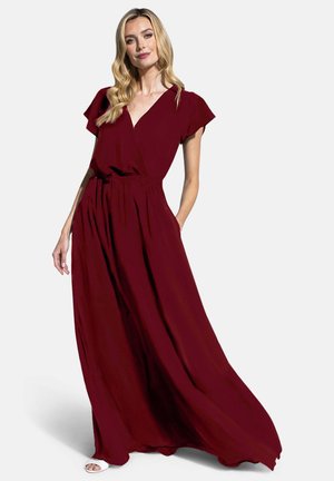 Žena s dlhými blond vlasmi, oblečená v splývavej hlboko červenej maxišatách s krátkymi rukávmi a bielych sandáloch s otvorenou špičkou, stojaca s jednou rukou v vrecku.