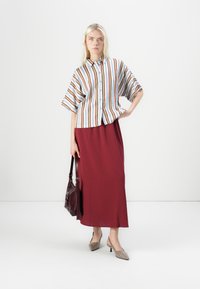 Camicia a righe blu, rossa e arancione con maniche larghe, abbinata a una lunga gonna bordeaux. La modella porta una borsa marrone e indossa tacchi grigi.
