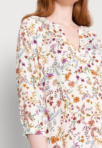 Bluse mit Blumenmuster, die einen cremigen Hintergrund, lebendige mehrfarbige Stickereien, einen V-Ausschnitt und Dreiviertelärmel mit gerafften Akzenten aufweist.