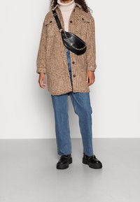 Abrigo de cuadros marrón de gran tamaño con grandes bolsillos, suéter beige de cuello alto, jeans azul claro y botas negras gruesas con acentos de cadena plateada.