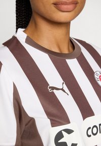Brown and white striped athletic jersey with a round neckline, featuring a small Puma logo and a large sponsorship patch. Textured material.