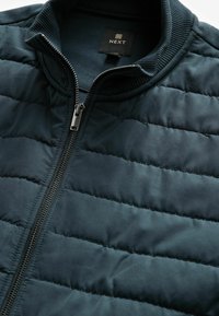 Veste matelassée vert foncé avec un col haut, fermeture éclair intégrale à l'avant et accents côtelés. Présente une texture lisse et légèrement brillante. Étiquette de la marque visible.