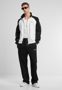 Sort og hvid zip-up jakke med sidepaneler, parret med sorte sweats, der har et broderet logo, og hvide sneakers.