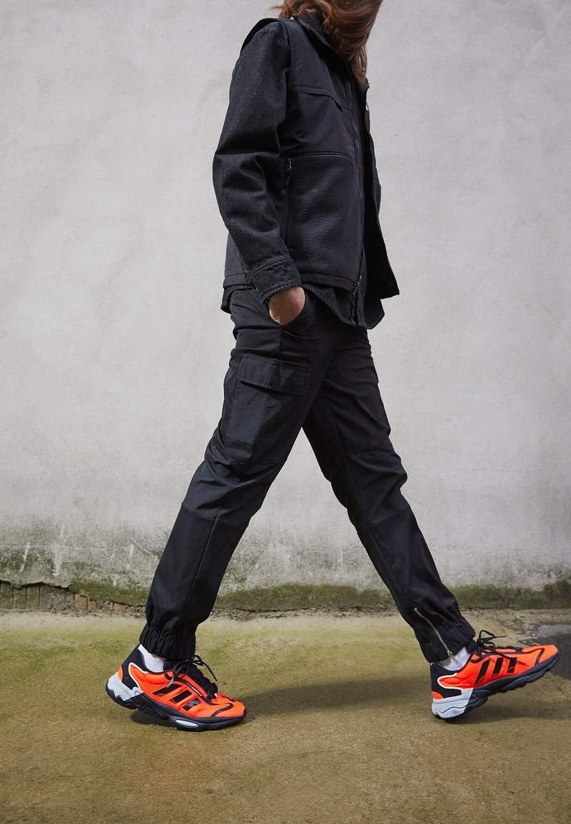 Černá bunda s fleecovou podšívkou, nákladní kalhoty a jasně oranžové tenisky s černými detaily. Bunda má zapínání na zip a elastické manžety.