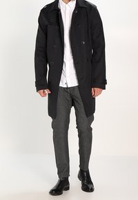 Svart trenchcoat med dubbelknäppt design, stora knappar, sidofickor, bärs över en vit skjorta och mönstrade svart-vita byxor.