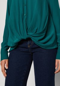 Blouse bleu canard à coupe ample, avec un devant boutonné et un détail en nœud à l'ourlet, associée à un jean bleu foncé avec des surpiqûres jaunes.