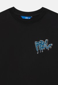Fekete crewneck póló kicsi kék graffiti-stílusú "REBEL" logóval, amely csepegő festék formájában helyezkedik el a bal mellen.