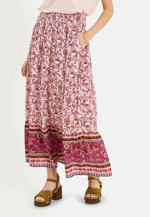 Mujer que lleva una falda maxi rosa con estampado floral y un borde ancho con patrón, y sandalias de plataforma marrones, con una mano en el bolsillo.
