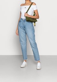 White off-the-shoulder cropped top with logo, high-waisted light blue jeans, and white sneakers. Green bag with adjustable strap.