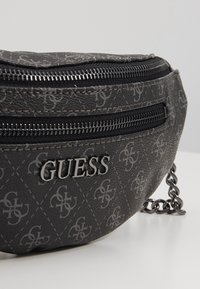 Sac banane noir Guess avec logo argenté, texture à motifs, deux compartiments zippés et détail de chaîne sur une surface blanche.