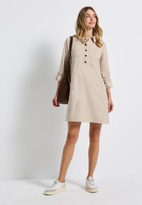 Robe beige à boutons en tissu léger. Elle présente un col, des manches longues, deux poches poitrine et une silhouette droite.