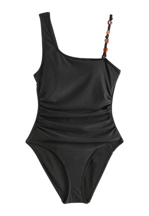 Žena s dlhými tmavými vlasmi oblečená v čiernych jednodielnych plavkách s jedným ramienkom s korálkovým pásikom, stojaca bosá vnútri pri bielej stoličke na slnečnom svetle.