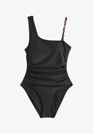 Hosszú sötét hajú nő fekete, egyrészes, egylábas fürdőruhában, gyöngyös pánttal, mezítláb áll egy fehér szék mellett beltéren napsütésben.