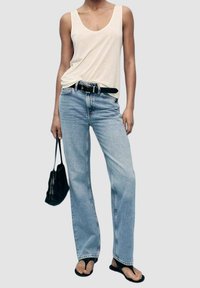 Personne portant un haut blanc sans manches, un jean taille haute bleu clair avec une ceinture noire, des tongs noires, et tenant un sac à main noir.