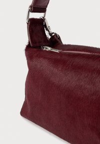 Malá obdélníková kabelka z bordó kožešiny se stříbrným zipem a nastavitelným ramenním popruhem na bílém pozadí.