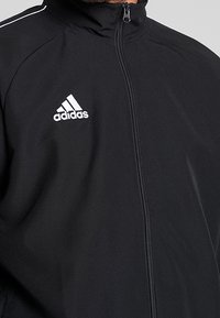 Svart zip-up jacka i lätt tyg. Har en hög krage, vit Adidas-logga på bröstet och subtila vita accentstänger.