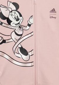 Giacca zip-up rosa chiaro con grafica in bianco e nero di un personaggio con un fiocco, decorata con nastri argentati e branding Adidas.