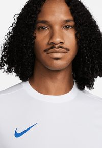 Maglietta atletica bianca con scollatura a girocollo e maniche corte. Presenta un logo Nike blu nella zona sinistra del petto. Tessuto testurizzato con finitura liscia.