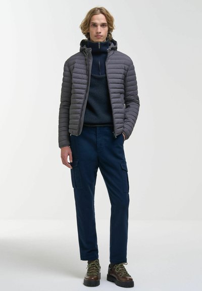 Jeune homme portant une doudoune grise à capuche, un pull bleu marine, un pantalon cargo bleu marine et des chaussures de randonnée marron, debout devant un fond clair uni.