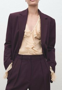 Blazer burdeos sobre una blusa crema con volantes. El blazer tiene un corte ajustado, mientras que la blusa presenta capas delicadas y una apariencia texturizada.
