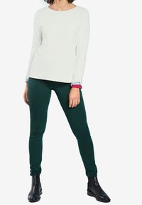 Haut long à manches blanches texturé avec un col rond et des accents de poignets roses, associé à un pantalon vert ajusté et des bottes-noires à la cheville.