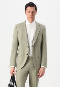 Traje ajustado de color verde claro con chaqueta de un solo botón, botones negros y bolsillos laterales. Llevado sobre una camisa blanca, la persona sostiene un bolso negro.