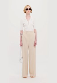 Top bianco a maniche corte con dettaglio allacciato, abbinato a pantaloni beige a gamba larga. Gli accessori includono occhiali da sole eleganti e una piccola borsa bianca.